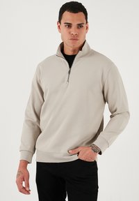 Béžový pulóver s vysokým golierom a polovičným zipsom, vyrobený z textúrovanej látky a s dlhými rukávmi. Spárovaný s čiernymi nohavicami s voľným strihom.