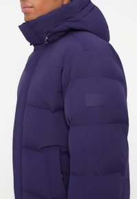 Man wearing a dark purple padded jacket with a high collar, visible side pocket, and a patch on the upper sleeve.