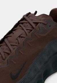 Gros plan sur une basket Nike marron avec une semelle noire texturée et un design à lacets sur un fond clair uni.