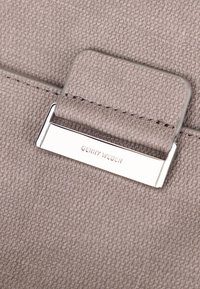 Beżowa torebka o fakturze z gładkimi srebrnymi akcentami, wyposażona w zamek z wygrawerowanym napisem "GERRY WEBER". Zbliżenie na materiał.