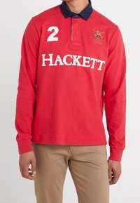 Polo-shirt rouge à manches longues avec col navy, logo "HACKETT" blanc et le numéro "2" sur le devant ; présente une texture unie et un design décontracté.
