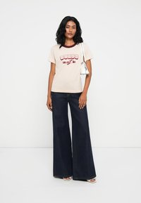 Μπεζ T-shirt με κόκκινο γραφικό "GUESS 1981" και μπορντό λαιμόκοψη, συνδυασμένο με σκούρα μπλε φαρδιά τζιν παντελόνια και ασημένιες ανοιχτές σανδάλες.