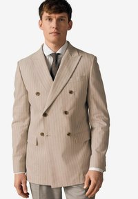 Double-breasted beige blazer with vertical stripes, notched lapels, and two front pockets. Paired with a white shirt and patterned tie.