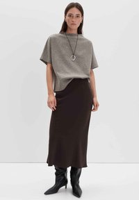 Eine Frau trägt ein lockeres graues Kurzarmoberteil, einen dunkelbraunen Midirock, schwarze spitze Stiefel und eine kreisförmige Anhängerkette vor einem schlichten Hintergrund.