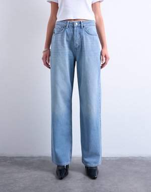 Osoba ubrana w jasnoniebieskie szerokie spodnie z wysokim stanem, biały krótki top, srebrne bransoletki i czarne buty na spiczastym nosku, stojąca wewnątrz pomieszczenia.