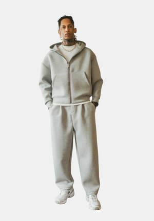 Homme debout les mains dans les poches, portant un sweat à capuche zippé gris, un pantalon de survêtement assorti, des baskets blanches et une chemise blanche sur un fond clair.