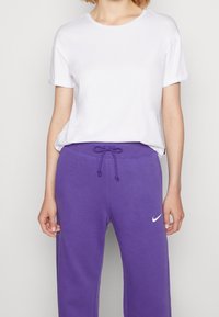 Vit t-shirt med kort ärm som är ihop med lila sweatpants med dragsnöre i midjan och en vit Nike-logotyp på vänster lår.
