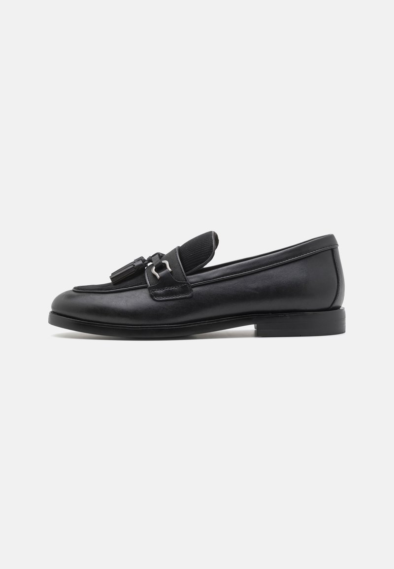 Mocassin en cuir noir avec des pompons et un petit talon, vu de profil sur fond blanc.