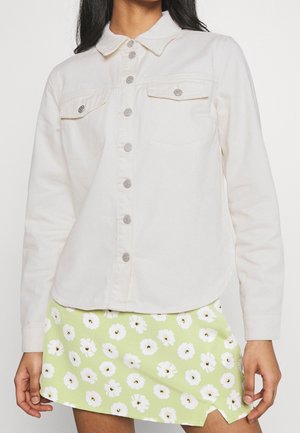 Femme portant une chemise blanche à manches longues boutonnée et une mini-jupe vert clair avec des motifs de fleurs blanches.