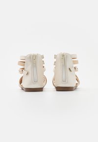 Beige sandaler med et glat materiale, med en baglynlås, flere stropper og en flad brun sål. Åben design for åndbarhed.