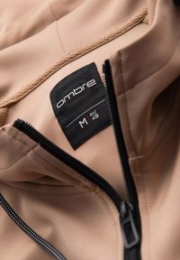Tessuto beige della giacca con una texture liscia; etichetta nera che mostra il marchio "ombre", taglia M e taglia europea 48. Presenta una chiusura a zip.