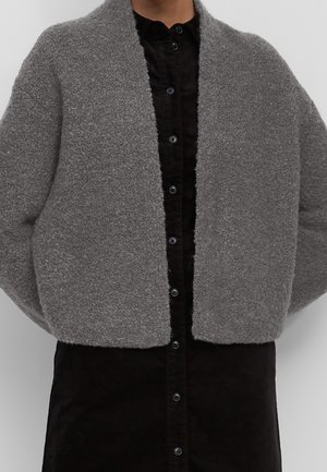 Osoba ubrana w krótki, teksturowany, szary wełniany żakiet na czarnej sukience koszulowej na guziki na jednolitym tle.