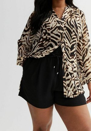 Frau trägt schwarze Shorts mit Kordelzug und eine langärmelige Bluse mit Zebramuster sowie mehrere goldene Ketten vor einfarbigem Hintergrund.