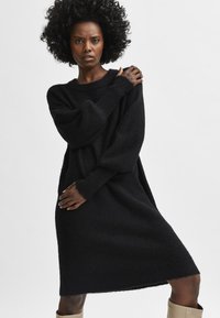 Juoda perdidinta megzta suknelė iš minkštos, tekstūruotos medžiagos. Turi ilgus rankovus, ribotas rankogalius ir laisvą siluetą.