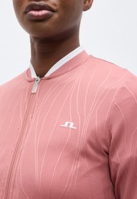 Κλείσιμο εικόνας ενός ατόμου που φοράει ροζ μπλούζα με φερμουάρ, με διακριτικές κάθετες υφασμάτινες γραμμές και ένα μικρό λευκό λογότυπο στο στήθος.