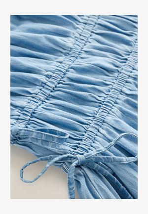 Šviesiai mėlynos džinsinės medžiagos su susiraukšlėta tekstūra. Centriniame siūle yra atvartai, o vienoje pusėje rišimui skirti raiščiai, rodo stengtą dizainą.