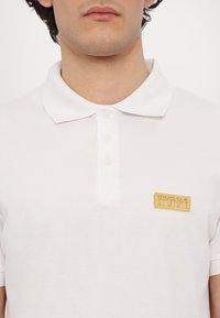 Bela polo majica iz teksturiranega materiala, s tremi sprednjimi gumbi in zlatim znakom "Versace Jeans Couture".