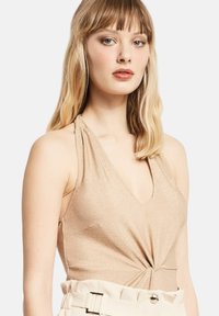 Top halter metallico oro con scollo a V e dettaglio frontale attorcigliato, realizzato in un tessuto lucido, abbinato a pantaloni beige a vita alta.