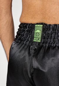 Melni satīna šorti ar elastīgu jostasvietu, uz kuras atrodas zaļa uzlīme ar uzrakstu "VENUM" un "RAŽOTS TAIZEMĒ" ar čūskas logotipu.