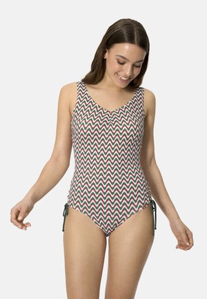 Mujer con un traje de baño de una pieza sin mangas con un patrón de zigzag en verde, rojo y blanco, de pie y mirando hacia abajo con una sonrisa.