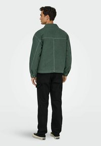 Chaqueta de mezclilla verde con costuras visibles, ajuste oversize y botones de presión, combinada con pantalones negros y zapatillas. Vista trasera.