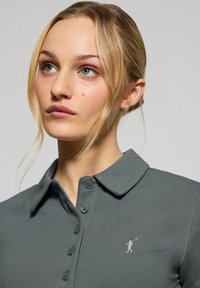 Mörkgrå polo skjorta med krage, med en knappslå och en liten broderad logotyp på vänster sida. Slät textur.