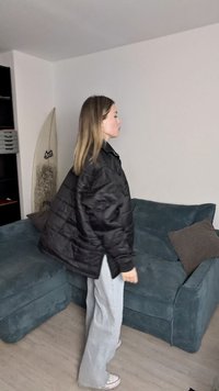 Μαύρο μπουφάν με κουκούλα και σχέδιο καπιτονέ, υπερμεγέθη εφαρμογή και ψηλό γιακά. Φορεμένο πάνω από ανοιχτό μπλε παντελόνι σε φαρδιά γραμμή και λευκά αθλητικά παπούτσια.