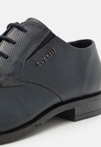 Chaussure en cuir gris foncé avec une finition texturée, présentant un design cousu et un petit logo en relief. Semelle en caoutchouc noir avec un talon bas.