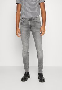 Hombre vistiendo jeans ajustados grises, botas negras hasta el tobillo y una camisa de manga corta a rayas en blanco y negro, de pie frente a un fondo blanco liso.