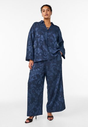 Žena oblečená v námornícky modrých širokých nohaviciach a zodpovedajúcom dlhom rukáve s kvetinovým žakárovým vzorom, stojaca s jednou rukou v vrecku.