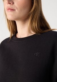 Čierna mikina z bavlnenej zmesi, s okrúhlym výstrihom a vyšitým logom 'cK' v zhodnej farbe na hrudi. Hladká textúra.