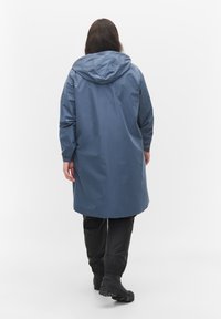 Imperméable bleu avec capuche, manches longues et coupe décontractée, associé à un pantalon noir ajusté et des bottes noires robustes.
