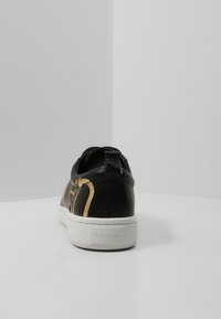Černé a zlaté tenisky s bílou podrážkou z pohledu ze zadní strany, s logem Calvin Klein na patě, na čistém bílém pozadí.