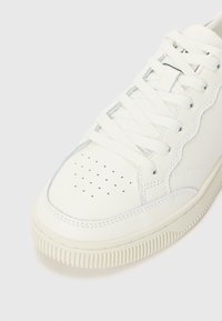 Hvite lær sneakers med en teksturert gummisåle, perforert tåhette, flate snører og et bølgete design langs sidene.