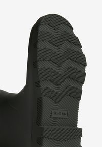 Semelle de botte en caoutchouc noir présentant un motif zigzag texturé pour une meilleure adhérence, avec un logo embossé sur le côté. Matériau supérieur lisse.