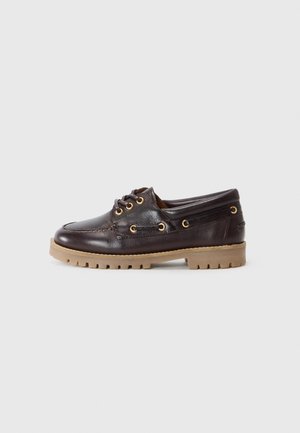 Chaussure bateau en cuir marron foncé avec un bout rond, œillets dorés et une semelle en caoutchouc texturée marron clair. Présente des détails de couture contrastants.