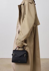 Borsa nera con texture e accessori dorati, presenta una patta triangolare e una maniglia corta. Indossata da una figura in un trench beige.