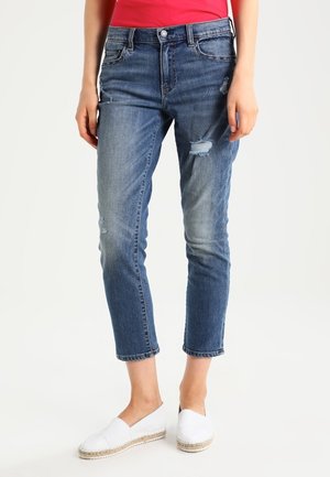 Mujer con vaqueros azules desgastados y recortados y zapatos blancos tipo espadrilles sin cordones, de pie frente a un fondo claro y liso.