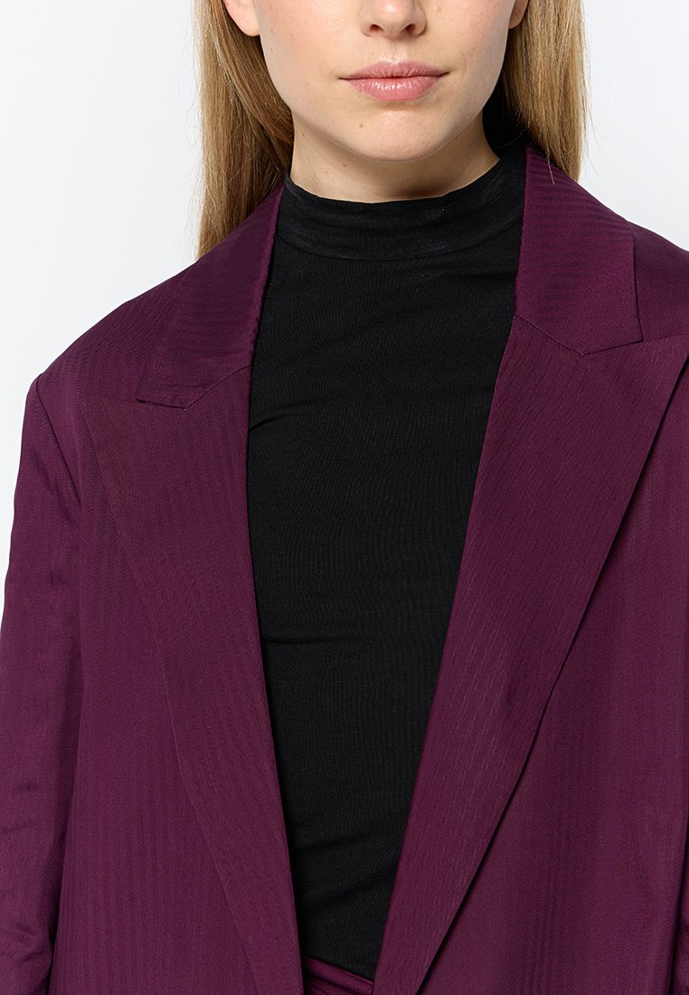 Blazer violet avec un tissu texturé, des revers nets et une silhouette ajustée, porté par-dessus un haut noir en col roulé côtelé.