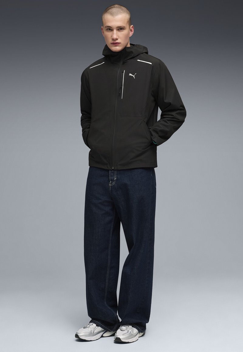 Jeune homme portant une veste à capuche noire Puma, un jean large foncé et des baskets argentées, debout les mains dans les poches devant un fond gris.