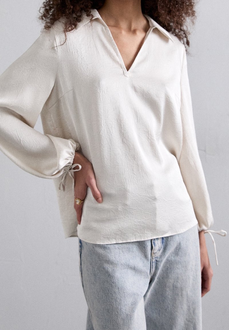 Blusa di colore crema con finitura testurizzata, scollatura a V e maniche lunghe con polsini elasticizzati. Abbinata a jeans di denim azzurri.