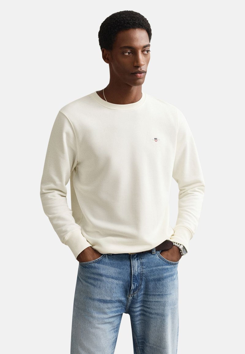 Ung mand med hænderne i lommerne iført en hvid langærmet sweater og lyseblå jeans, der kigger let mod højre.