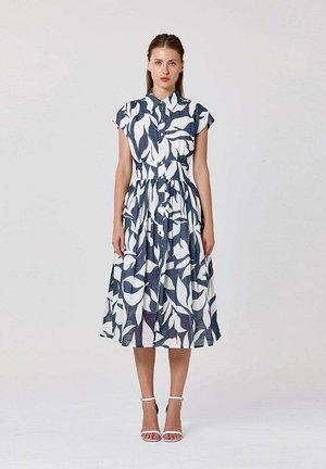 Femme debout portant une robe midi sans manches à boutons avec un grand imprimé feuilles bleu marine et blanc et des talons blancs à bride à la cheville sur un fond uni.