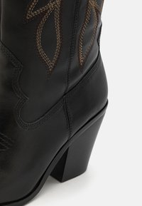 Bota de vaquero de cuero negro con costuras decorativas en marrón, puntera puntiaguda y un alto tacón apilado. Presenta bordes superiores curvados y una textura sutil.