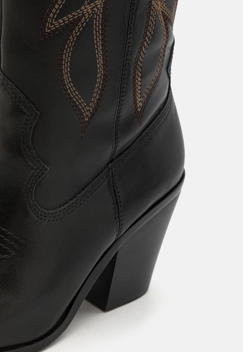 Stivale da cowboy in pelle nera con decorazioni in cucitura marrone, punta affusolata e tacco alto impilato. Presenta bordi superiori curvi e una texture sottile.