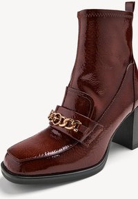 Bottine en cuir verni marron avec un bout carré, un talon bloc et un ornement de chaîne dorée sur la bride avant ; texture lisse et finition brillante.
