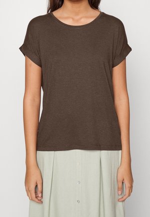 Kvinde iført en kortærmet mørkebrun t-shirt med opbukket kant og en lys beige nederdel med knapper, stående mod en ensfarvet baggrund.