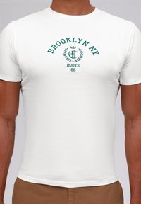 Camiseta blanca de manga corta con "Brooklyn NY" y "Route 66" en texto verde, con un gráfico de laurel sobre el texto. Textura de tela suave.