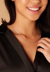 Lachende vrouw met een dunne zilveren ketting en een zwarte satijnen top, die met haar rechterhand haar decolleté aanraakt.