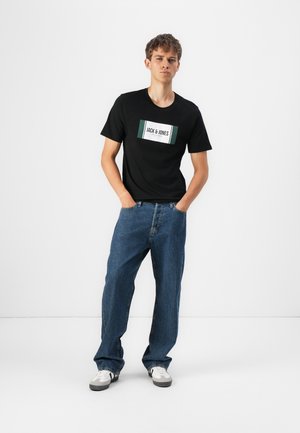 Svart kortermet t-skjorte med en hvit og grønn logo, kombinert med løse blå denimjeans og hvite joggesko. Enkel hvit bakgrunn.